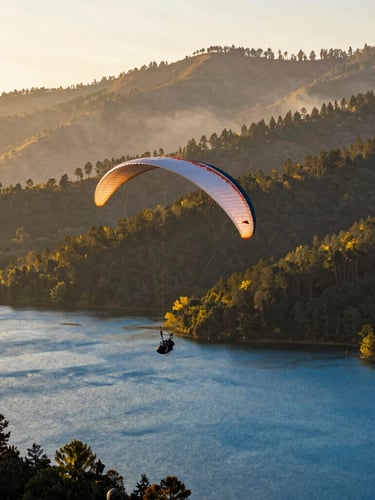 Wide environmental shot of a single paraglider soaring silently over a deep blue lake in Valle de Bravo, warm golden hour sunlight reflecting off the water, mist rising from the distant pine-covered mountains.