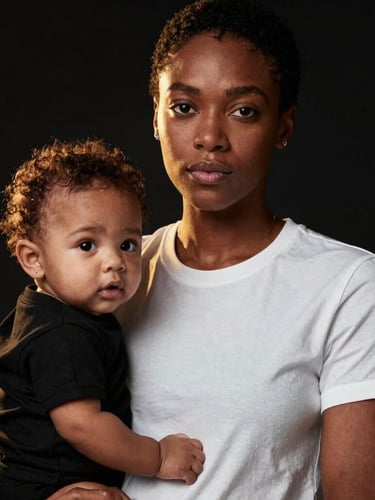 Medium close-up of a proud Black mother holding her baby, both wearing minimalist black and white statement tees, high-contrast dramatic studio lighting, warm gold backlighting, deep black background.