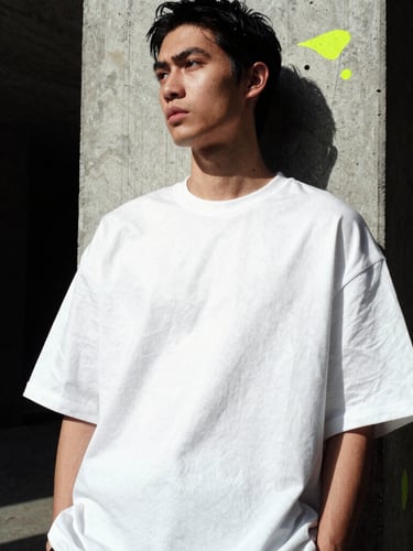 Wide architectural crop of a model leaning against a raw concrete pillar, wearing an oversized white drop-shoulder tee with heavy fabric folds, dramatic side-lighting.