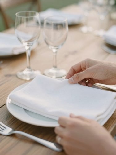 Close-up of hands arranging delicate white linen napkins and elegant glassware on a rustic wooden table, soft diffused daylight, shallow depth of field.