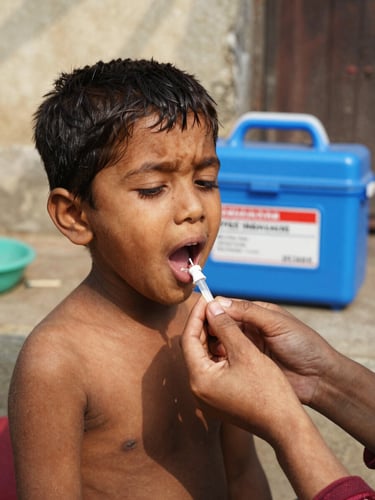 A close-up documentary photograph of a child receiving oral polio drops under warm, natural sunlight, with the iconic blue vaccine carrier box visible in the soft-focus background.
