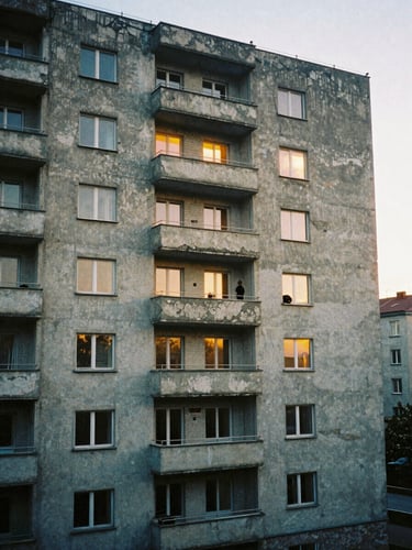 Portrait-format film poster composition, crumbling brutalist apartment block at twilight, warm amber windows punctuating a grey concrete facade, a single figure on a high balcony, Eastern European urban mood