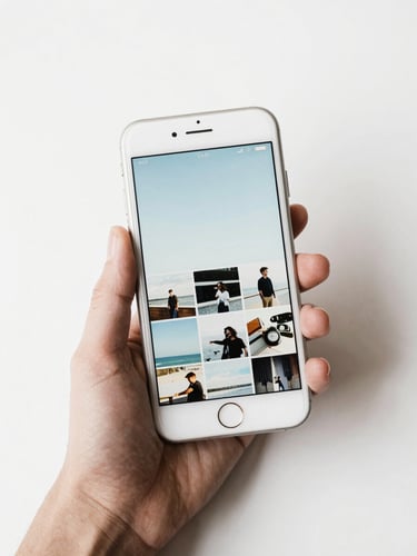 Close overhead flatlay of a hand holding a white iPhone displaying a bright photo gallery app interface with soft blue tones, clean white background beneath, natural daylight, tight framing on the device screen