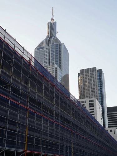 Low-angle architectural perspective of structural scaffolding lines against the Melbourne skyline, crisp morning daylight, desaturated navy and concrete tones, sharp red accents, wide-angle lens