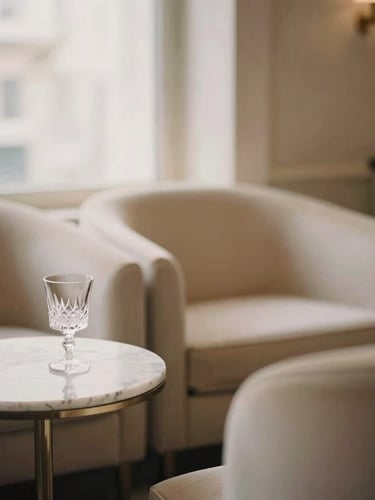 Editorial portrait of a sophisticated private VIP lounge with minimalist champagne-colored furniture, a marble side table with a crystal glass, soft warm window light, elegant atmosphere, shallow depth of field.