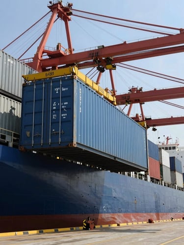 Industrial shipping container being loaded onto a cargo vessel at a bustling harbor, bright daylight, high-contrast maritime setting.