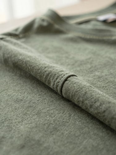 Macro close-up of perfectly intact double-stitched seams on a well-worn sage green cotton t-shirt, showing tactile fabric texture, soft morning side-light, shallow depth of field