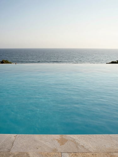 Overhead architectural view of a rectangular infinity pool edge meeting the Atlantic horizon, midday light, water surface perfectly still, pool coping stones in pale limestone, sea visible beyond the horizon line, no people