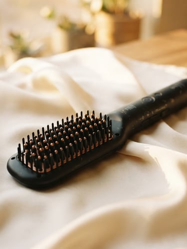 Elegant close-up of a premium matte black hair straightening brush resting on a soft cream-colored silk fabric, warm golden-hour window light, shallow depth of field, 35mm