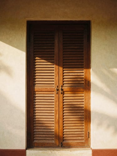 A tall portrait of a louvered wooden screen in a Goan villa, light slicing through to create dramatic shadows on lime-plaster, medium format film.