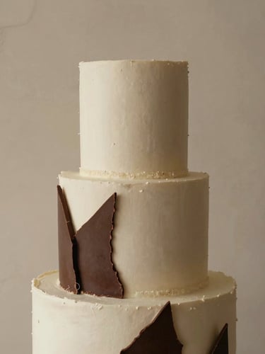 Elegant editorial food photography, soft natural side-light, close-up of a minimalist sculptural wedding cake with delicate chocolate shards, set against a textured plaster backdrop, warm tones.