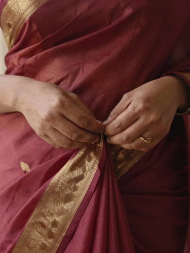 Close-up of hands meticulously pleating a rich crimson and gold silk saree, warm diffused lighting, editorial aesthetic, 35mm