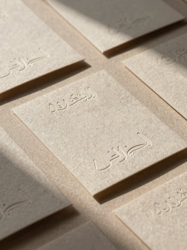 A cinematic, wide 21:9 frame of tactile brand collateral, textured linen paper packaging with elegant Arabic embossing, warm afternoon sunlight casting soft organic shadows across a neutral sand surface.