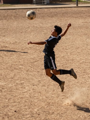 Low-angle heroic shot of a young player mid-air striking the ball on a dirt pitch, harsh midday sun casting deep shadows, raw high-stakes athletic intensity, gritty sports photography.