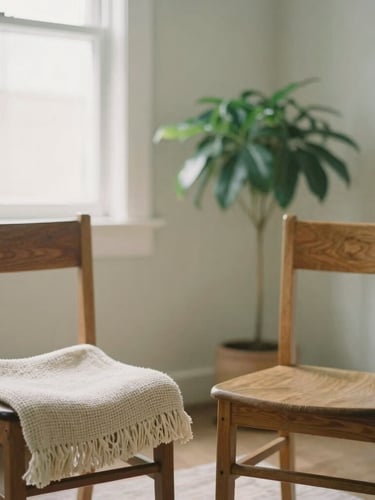 Medium portrait shot of a comfortable therapy space, two empty wooden chairs facing each other with a soft woven throw blanket, a green potted plant in the background, soft natural window light, muted sage and cream tones, shallow depth of field.