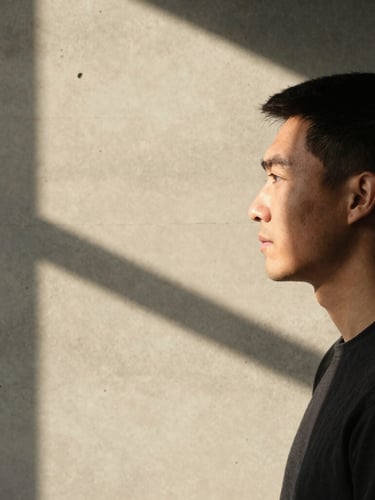 Portrait of a creative professional in profile, harsh direct sunlight casting a sharp shadow of window frames onto a warm concrete wall, minimalist composition, warm tones, subtle grain.