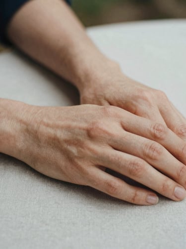 Extreme close-up of a person's hands resting quietly on a linen-covered table, soft diffused overcast daylight, visible film grain, warm tones.