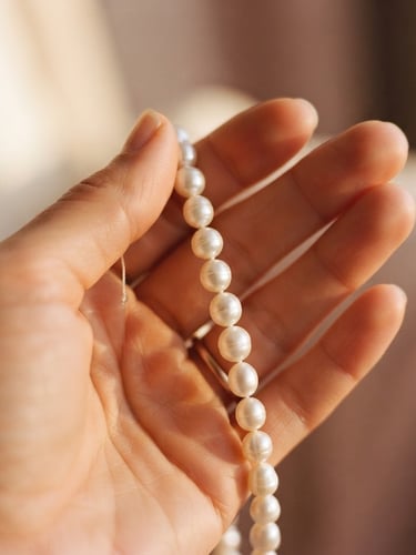 Macro shot of a hand delicately holding a strand of luminous freshwater pearls, warm golden-hour light, cream and dusty rose tones, shallow depth of field, worn on skin.
