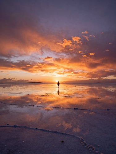 Cinematic wide shot of the Salar de Uyuni at sunset, deep perspective, dramatic orange and purple sky reflecting on the water-covered salt flats, a single silhouette of a traveler in the distance.