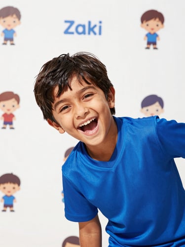 Bright commercial photography of a laughing South Asian child wearing a vibrant blue sports jersey, warm sunny daylight, playful energetic mood, white background with Zaki character stickers, crisp focus.