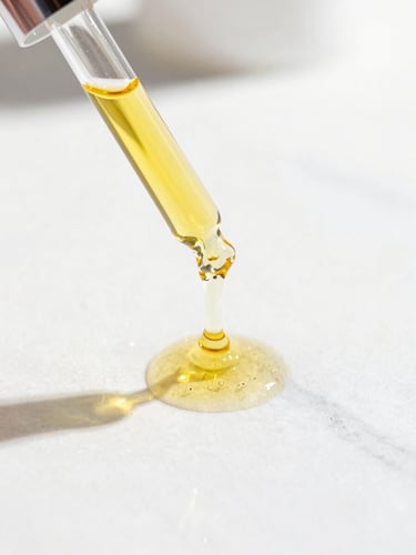 Bright editorial photography, macro details of liquid golden botanical oil dripping from a glass pipette onto a clean white marble surface, sun-drenched highlights, sharp focus, 35mm lens