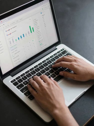 Overhead close-up of hands typing on a laptop keyboard, a browser tab showing a Google Analytics dashboard visible on screen, cool even studio lighting, minimal dark desk surface
