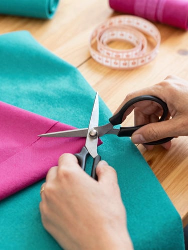 Close-up of hands carefully cutting colorful teal and magenta fabrics with metal scissors on a wooden table, soft measuring tape nearby, warm workshop light
