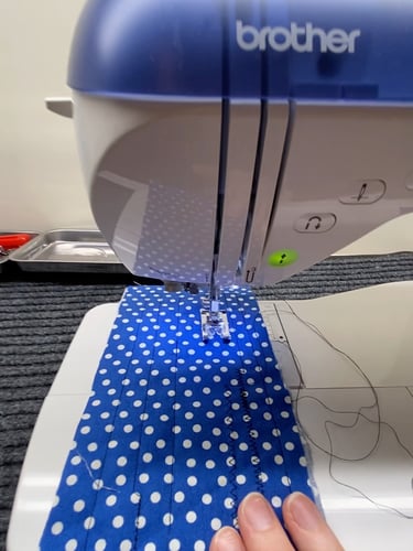 A hand feeding blue polka-dot fabric through the needle and foot area of a sewing machine.