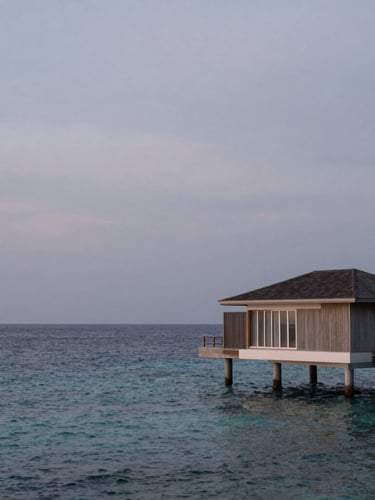 Extreme wide environmental shot of a single minimalist overwater villa at twilight, soft overcast light, endless calm ocean, negative space dominating the left side