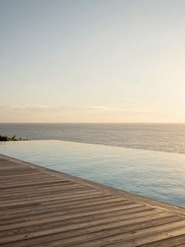 Extreme wide environmental shot of a private infinity pool merging with the open ocean at golden hour, minimalist wooden deck, warm low sunlight