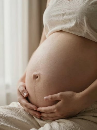 Close-up of gentle hands cradling a bare pregnant belly, warm diffused window light from the left, soft cream linen visible at the edge of the frame, intimate and still