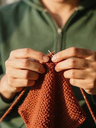 Close-up of hands knitting with textured terracotta wool, warm golden-hour sunlight filtering through window shadows, shallow depth of field, 35mm photography