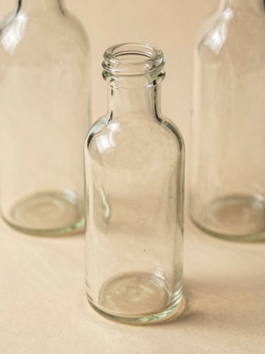 Macro close-up of clean glass bottles on a warm beige surface with soft morning shadows, minimal aesthetic.
