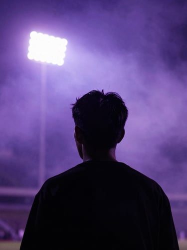 Aggressive close-up of a supporter's silhouette in a stadium filled with purple smoke, backlit by intense floodlights, raw urban night aesthetic