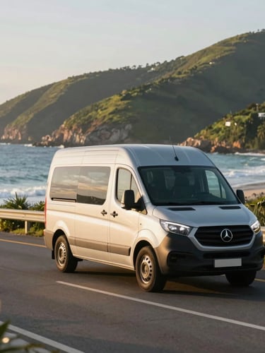 A modern silver passenger van driving along a scenic coastal road in Florianópolis, blue ocean and green hills in the background, warm golden hour light, 35mm