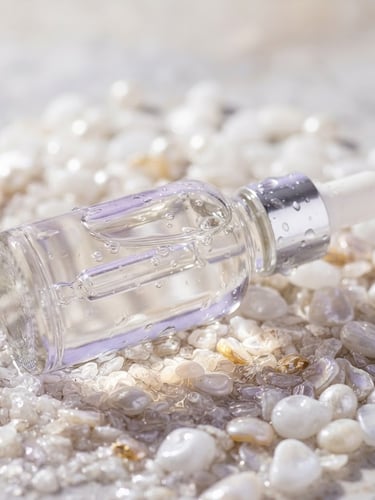 Macro shot of a clean glass dropper bottle reflecting soft lavender and gold light on a wet pearl stone surface, high-key studio lighting.