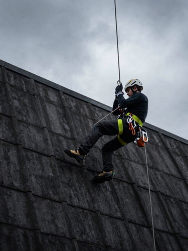Low-angle dynamic shot of a professional industrial climber in high-visibility safety harness suspended on ropes against a dark tiled roof, dramatic overcast sky, sharp focus on technical carabiners.