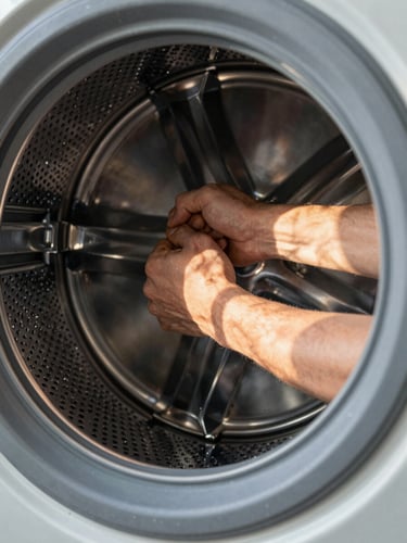 Extreme close-up, tight frame: a technician's hands gripping and pulling the inner drum of a front-load washing machine, fingers wrapped around the metal tub rim, workshop work-light casting hard shadows across the steel drum and lint filter housing, natural daylight entering from a nearby window illuminating scattered water droplets on the drum surface — no faces, no studio setup, no styling.