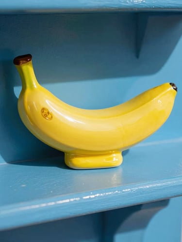 Close-up of a bright yellow ceramic banana sculpture on a cobalt blue mantelpiece, direct sunlight with sharp playful shadows, vibrant retro-pop aesthetic