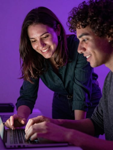 A supportive female mentor guiding an adult student typing on a laptop, warm purple ambient screen glow, natural smiles, close-up documentary style.