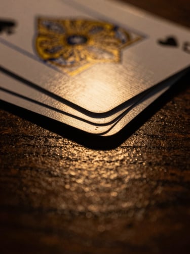 Close-up macro shot of elegant playing cards with gold-gilded edges, dramatic low-key lighting, deep shadows, warm gold light reflecting off a dark wooden surface.