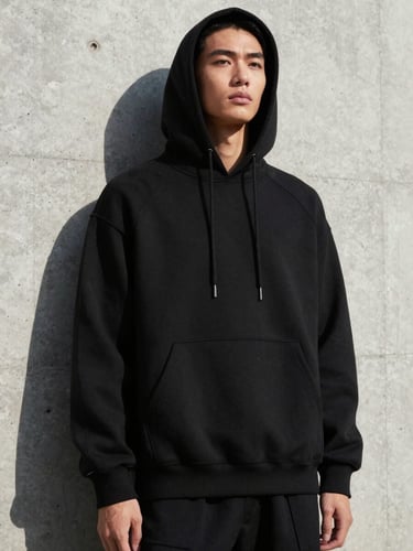 Low-angle editorial fashion shot of a model in a heavy black structured hoodie against a raw concrete wall, sharp direct sunlight, deep shadows, 35mm lens