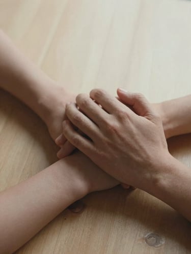 Close-up of two hands intertwined resting on a sunlit wooden surface, soft overcast diffused light, skin texture visible, warm sand and terracotta tones, intimate and unhurried mood