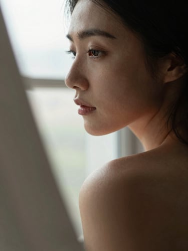 Close-up portrait frame, a woman's profile looking out a sun-drenched window, soft overcast daylight diffusing across her face and bare shoulder, warm skin tones, shallow depth of field, fabric texture visible in foreground