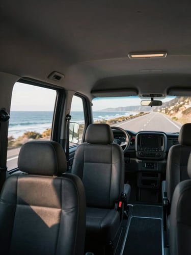 Interior of a clean modern van seen from the passenger perspective, rows of dark seats, window light streaming in from the left side, coastal road visible through the front windshield, natural daylight, no passengers