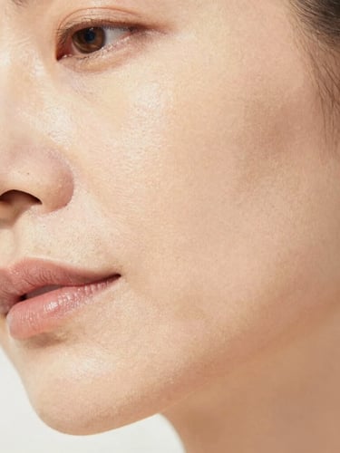 Close-up of a mature woman's cheek, soft natural daylight illuminating subtle skin texture, shallow depth of field, warm off-white tones, serene expression. No harsh lines or exaggerated features.