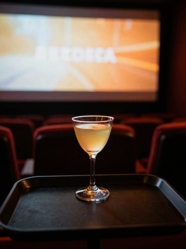 First-person perspective of a dark theater seat tray with a glowing craft cocktail, soft out-of-focus cinema screen in the background, neon-amber accent lighting, 35mm