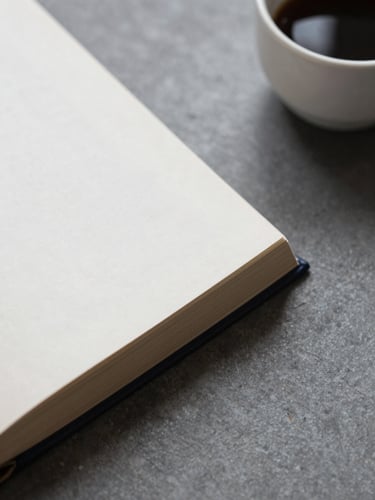 Overhead detail of a closed hardcover book beside a small coffee cup on a slate-grey surface, soft north light, minimal and still, no text visible