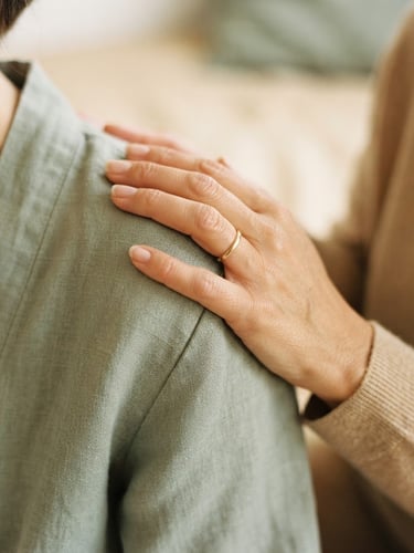 Extreme close-up of hands in a supportive, slow somatic touch on a shoulder, soft natural morning light, warm sand and muted sage tones, minimalist editorial photography, shallow depth of field, 35mm