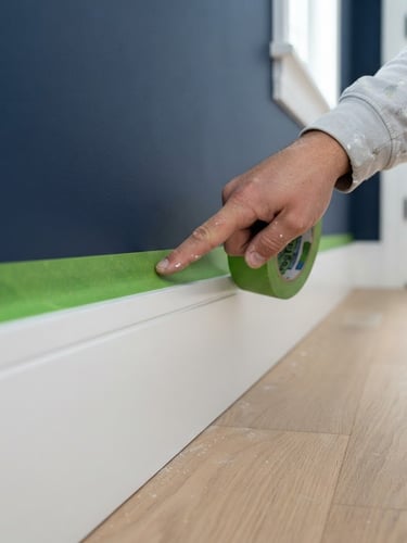 Macro close-up of painter applying crisp green painter tape to a pristine white baseboard, sharp paint lines, bright natural daylight, high-end residential interior